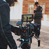 A filmmaker is operating a professional camera mounted on a tripod, capturing a scene from a film set that resembles a Western town. The camera's monitor displays an image of a person dressed in a period costume, suggesting the filming of a historical or Western-themed production. The person near the camera is dressed warmly, indicating cool weather.